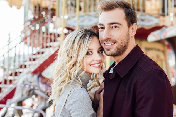 портрет улыбающейся влюбленной пары в осеннем наряде, смотрящей на камеру возле карусели в парке развлечений

