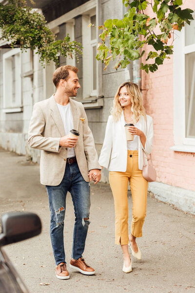 cheerful couple in autumn outfit holding hands, looking at each other and walking on street with disposable coffee cups