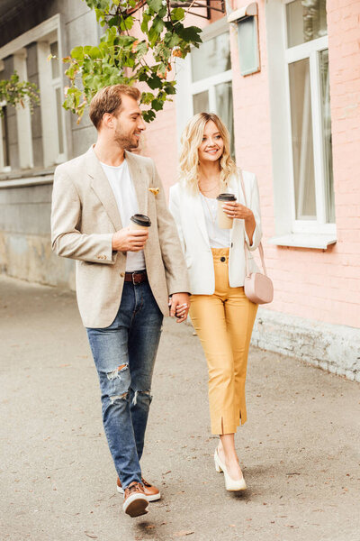 улыбающаяся пара в осеннем наряде, держащаяся за руки и идущая по улице с одноразовыми кофейными чашками

