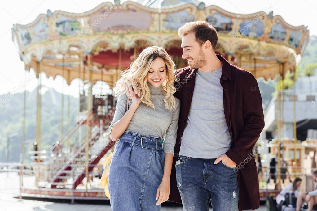 smiling couple in autumn outfit walking and hugging near carousel in amusement park