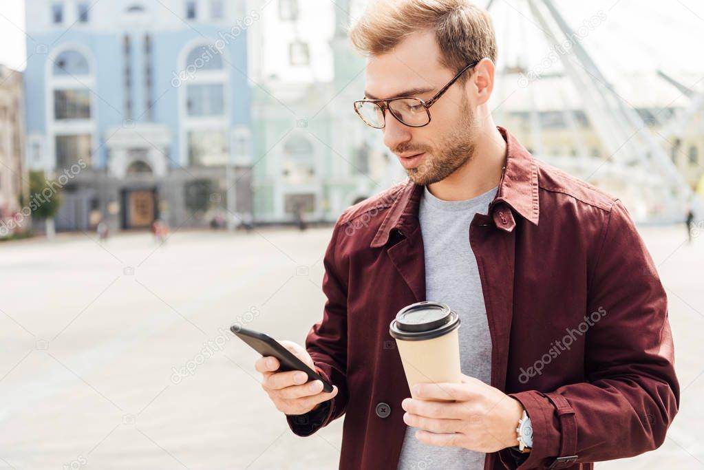 handsome man in autumn outfit holding coffee to go and using smartphone in city
