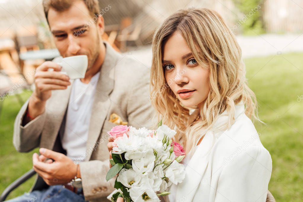 attractive girlfriend holding bouquet of roses in cafe, boyfriend drinking coffee