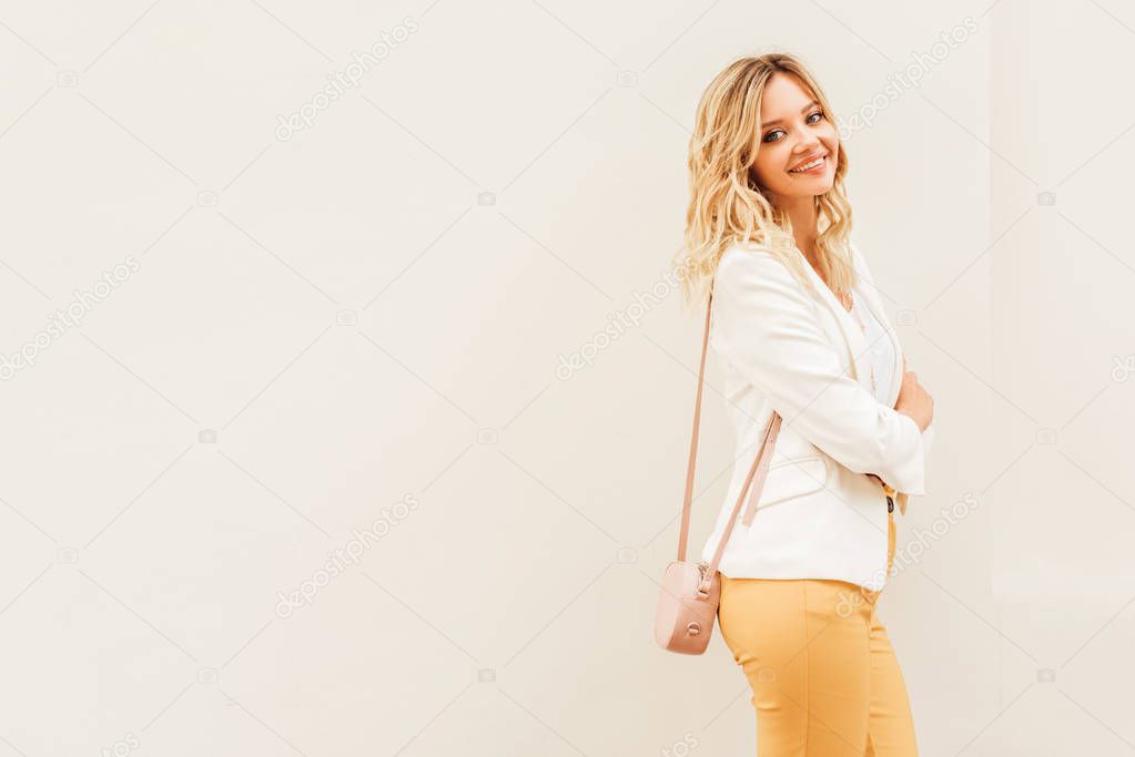 smiling beautiful stylish woman in autumn outfit standing near beige wall in city and looking at camera