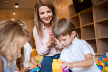 smiling educator looking at children playing with constructor in kindergarten clipart