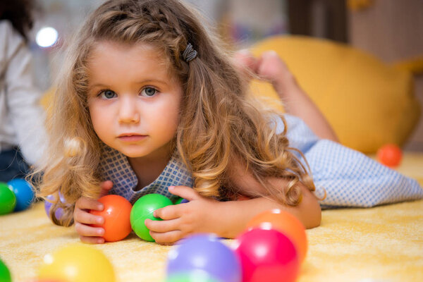 adorable child lying on carpet with colorful balls in kindergarten and looking at camera
