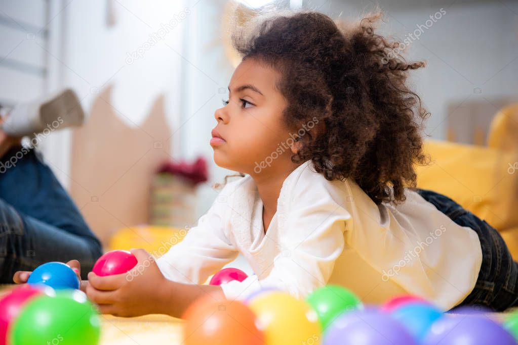 side view of adorable african american kid lying on carpet with toys and looking away in kindergarten
