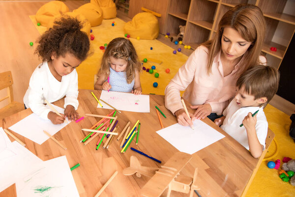 high angle view of tutor and multicultural children drawing together in kindergarten