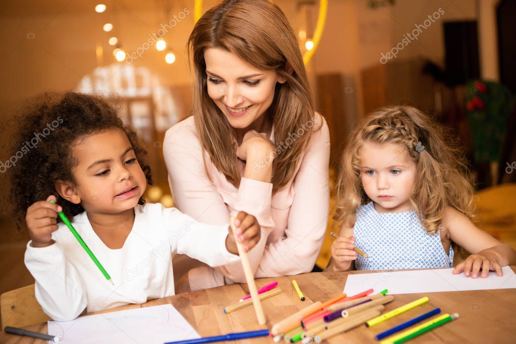smiling educator sitting near multiethnic children drawing in kindergarten