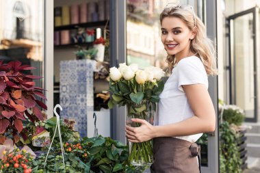 attractive florist holding jar with white roses near flower shop and looking at camera