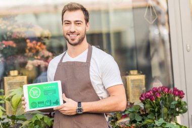 smiling handsome florist holding tablet with shopping appliance near flower shop clipart