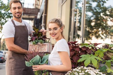 smiling female and male florists holding potted plants near flower shop and looking at camera clipart
