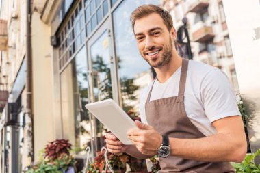 Tablet çiçek dükkan yakın tutarak ve kameraya bakarak yakışıklı çiçekçi gülümseyen