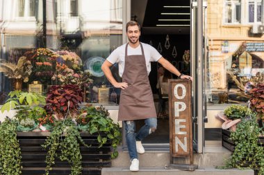 smiling handsome flower shop owner standing on stairs and leaning on open sign clipart