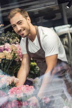buketler çiçek dükkanında bakımı ve kameraya bakarak gülümseyen yakışıklı çiçekçi, pencereden görüntüleme