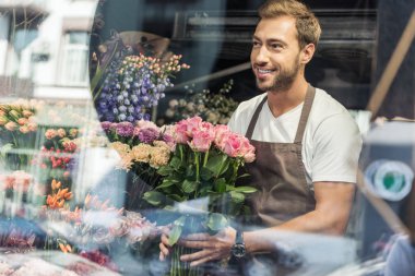 view through window of handsome florist holding bouquet of pink roses in flower shop and looking away clipart