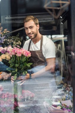 pembe gül buketi Çiçekçi dükkanı tutarak ve kameraya bakarak yakışıklı çiçekçi, pencereden görüntüleme