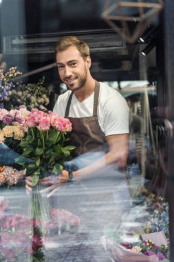 view through window of handsome florist holding bouquet of pink roses in flower shop and looking at camera clipart