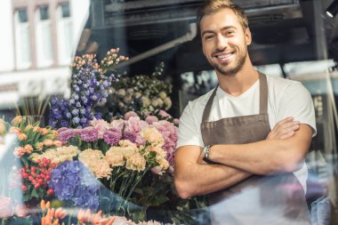 view through window of handsome florist standing with crossed arms in flower shop and looking at camera clipart