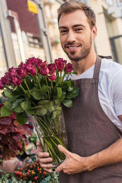 Улыбающийся красивый флорист держит стеклянную банку с бордовыми розами рядом с цветочным магазином
