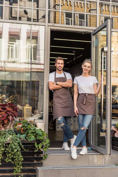 young smiling florists standing near flower shop and looking at camera