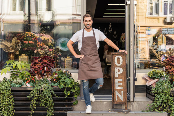 smiling handsome flower shop owner standing on stairs and leaning on open sign