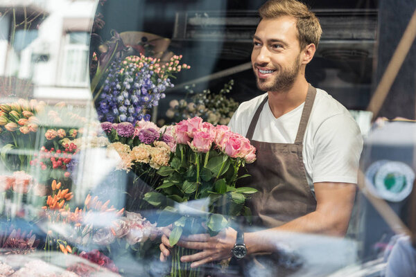view through window of handsome florist holding bouquet of pink roses in flower shop and looking away