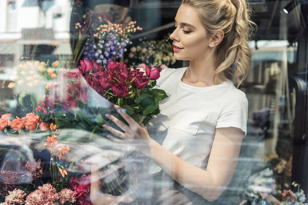 view through window of attractive florist holding bouquet of burgundy roses in flower shop