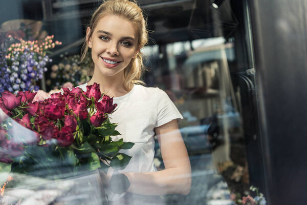 view through window of smiling attractive florist holding bouquet of burgundy roses in flower shop