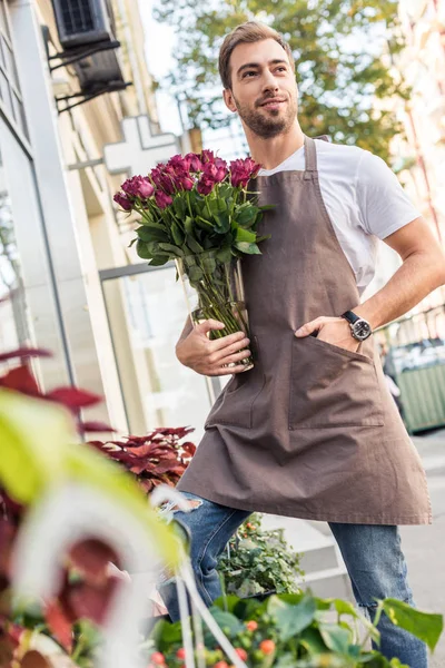 Low angle view of handsome florist holding glass jar with burgundy roses near flower shop and looking away — Stock Image Low Angle View Handsome Florist Holding Glass Jar Burgundy Roses Royalty Free Stock Photos