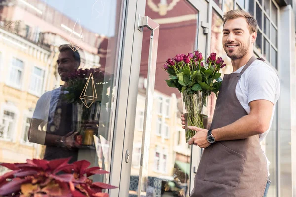 Low angle view of handsome florist holding glass jar with burgundy roses near flower shop and looking at camera — Stock Image Low Angle View Handsome Florist Holding Glass Jar Burgundy Roses Royalty Free Stock Images