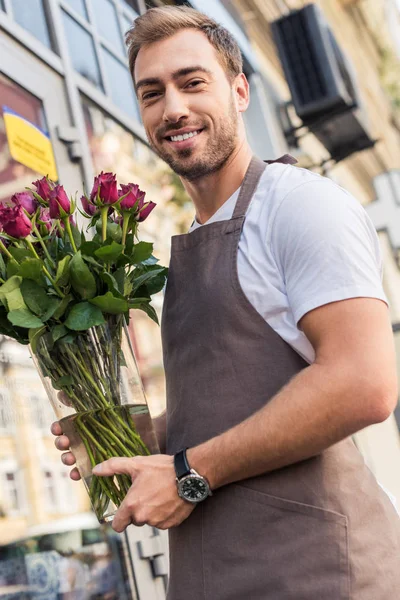 Handsome happy florist holding glass jar with burgundy roses near flower shop — Stock Image Handsome Happy Florist Holding Glass Jar Burgundy Roses Flower Shop Stock Image