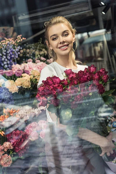 View through window of attractive florist holding bouquet of burgundy roses in flower shop and looking at camera — Stock Image View Window Attractive Florist Holding Bouquet Burgundy Roses Flower Shop Stock Photo