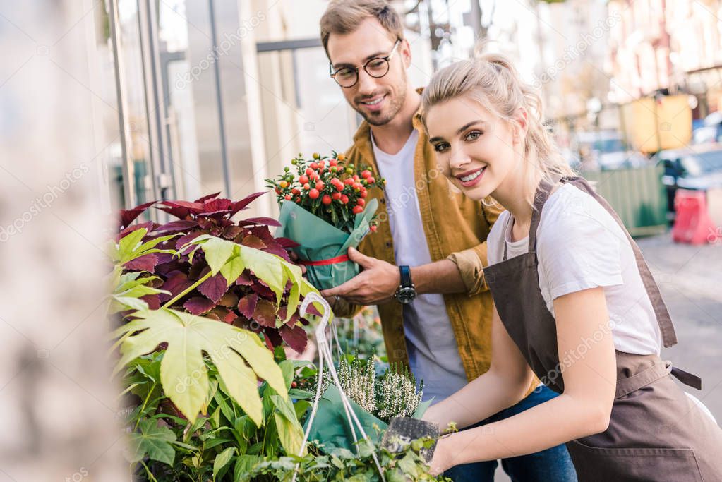 smiling florist and customer standing with plants near flower shop