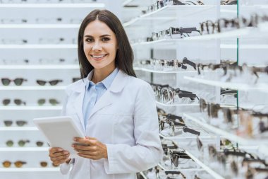 beautiful smiling optician using digital tablet near shelves with eyeglasses in ophthalmic shop clipart