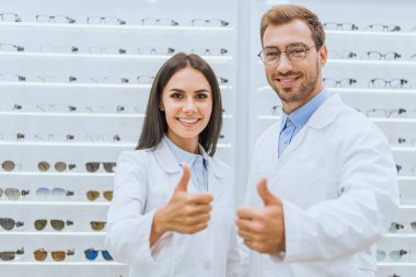 professional doctors in white coats showing thumbs up in optics with eyesight on shelves clipart