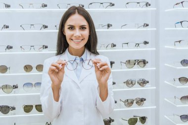 beautiful smiling optometrist holding eyeglasses in ophthalmic shop clipart