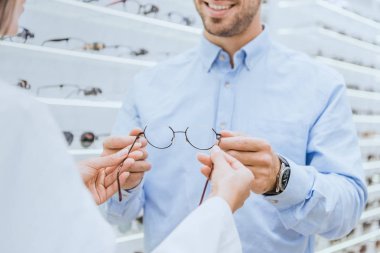 cropped image of female oculist giving eyeglasses to young man in ophthalmic shop clipart