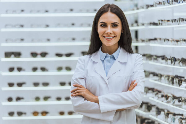 professional confident optician posing with crossed arms near shelves with eyeglasses in optica