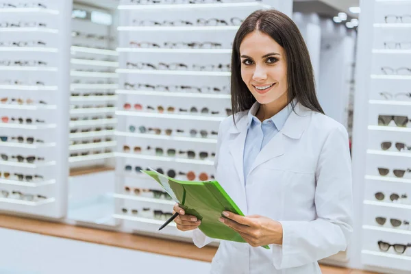 Hermosa Sonriente Oculista Sosteniendo Papeles Pluma Tienda Oftálmica —  Fotos de Stock