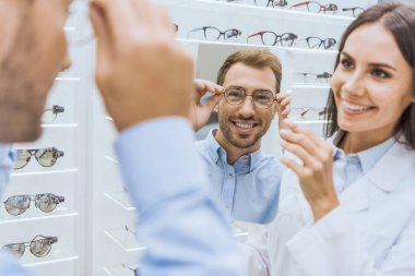 cheerful female optometrist holding mirror while smiling man choosing eyeglasses in ophthalmic shop clipart