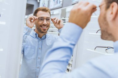 neşeli genç adam Gözlük seçimi ve optica aynaya bakarak kısmi görünümü 