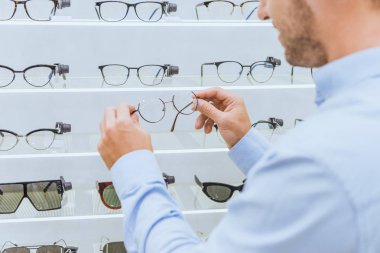 partial view of man taking eyeglasses from shelves in optics clipart