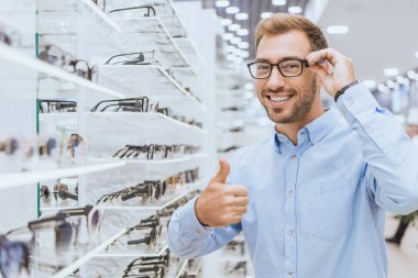 happy young man choosing eyeglasses and doing thumb up gesture in optica clipart