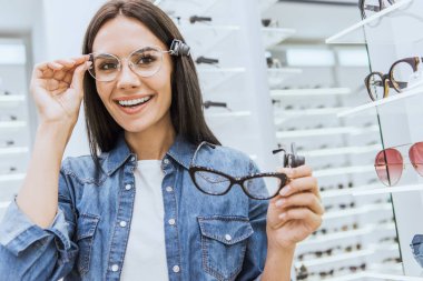 portrait of attractive happy woman choosing eyeglasses and looking at camera in ophthalmic shop