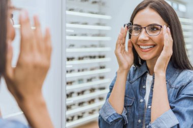 partial view of smiling woman choosing eyeglasses and looking at mirror in optica  clipart