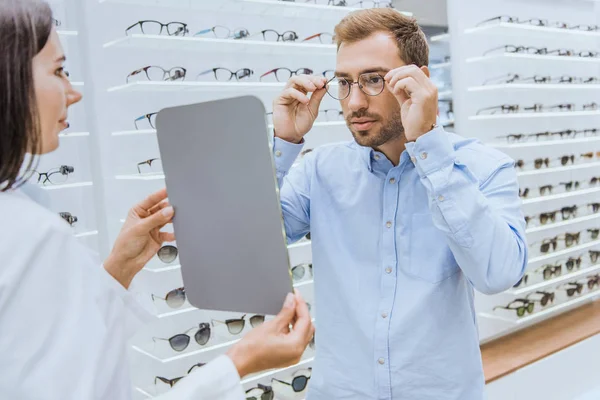 Visão Lateral Optometrista Feminino Segurando Espelho Enquanto Homem Escolhe Óculos — Fotos gratuitas