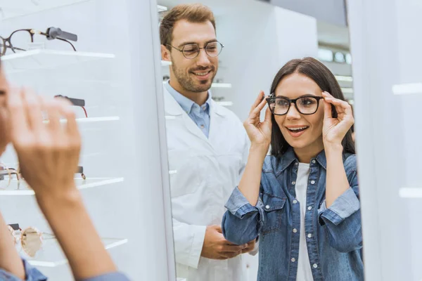 Attractive Smiling Woman Choosing Eyeglasses Looking Mirror While Male Optometrist — Stock Photo, Image