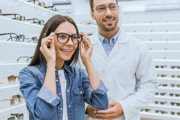 happy woman choosing eyeglasses while male oculist standing near in ophthalmic shop