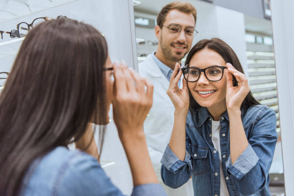 задний вид веселой женщины, выбирающей очки и смотрящей в зеркало, в то время как мужчина-оптометрист стоит рядом в оптике
 