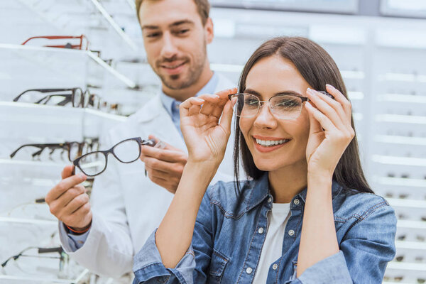 beautiful smiling woman choosing eyeglasses while male oculist standing near with another eyeglasses in ophthalmic shop